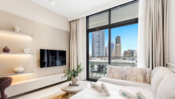 Ett modernt hotellrum med ett stort fönster, en platt-TV, en soffa, ett litet bord med en växt och en dekorativ hylla.