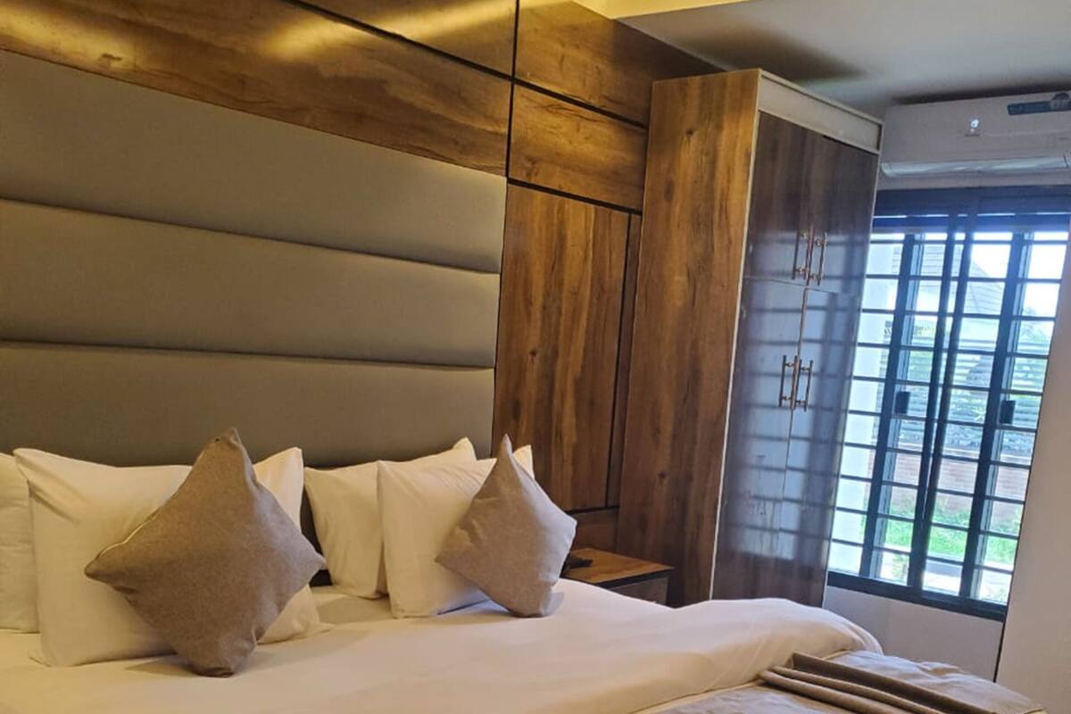 A large bed with multiple pillows, a wooden headboard, and a window with blinds.