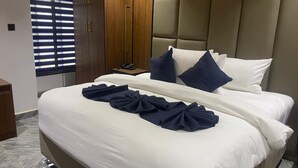 A hotel room with a large bed, a wooden wardrobe, a bedside table with a phone, and a window with blinds.