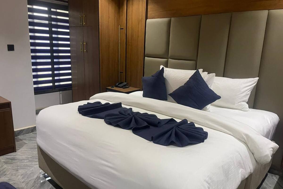 A hotel room with a large bed, a wooden wardrobe, a bedside table with a phone, and a window with blinds.