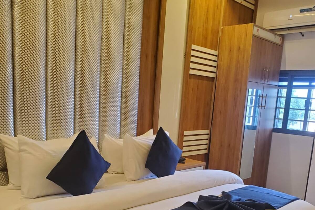 A double bed with white bedding and dark blue pillows, a wooden wardrobe, and a window with curtains.