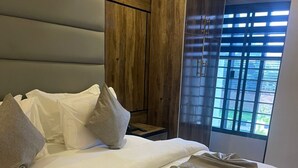 Ein ordentlich bezogenes Bett mit Kissen, ein Holzschrank und ein Fenster mit Jalousien.