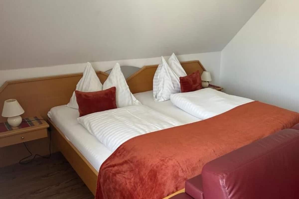 Una cama individual con cabecera de madera, ropa de cama blanca y detalles en rojo. Hay una mesita de noche con una lámpara y un enchufe. Un sofá de cuero rojo está ubicado al pie de la cama.