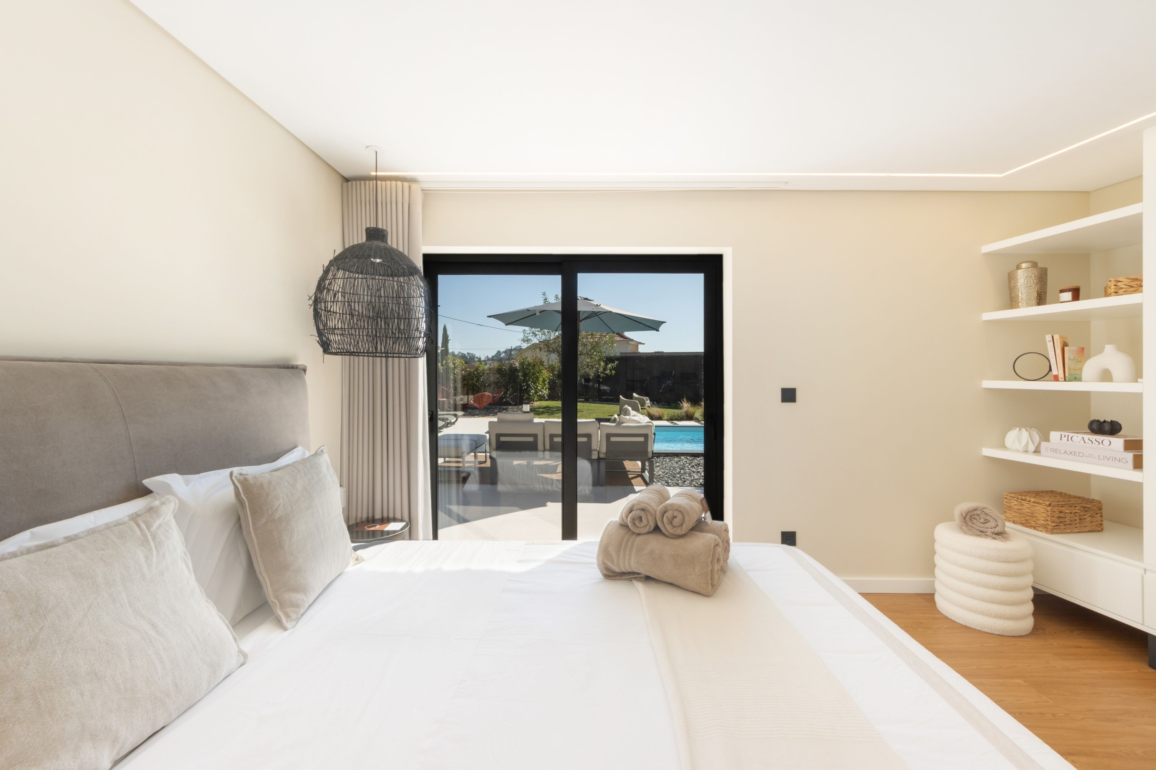 Ein modernes Schlafzimmer mit einem großen Bett, Blick auf ein Außenbecken und einer weißen Regaleinrichtung mit Dekoration.