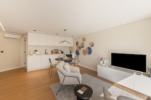 A modern living room with a flat-screen TV, a dining area with a table and chairs, and a kitchen with white cabinets and a built-in oven.