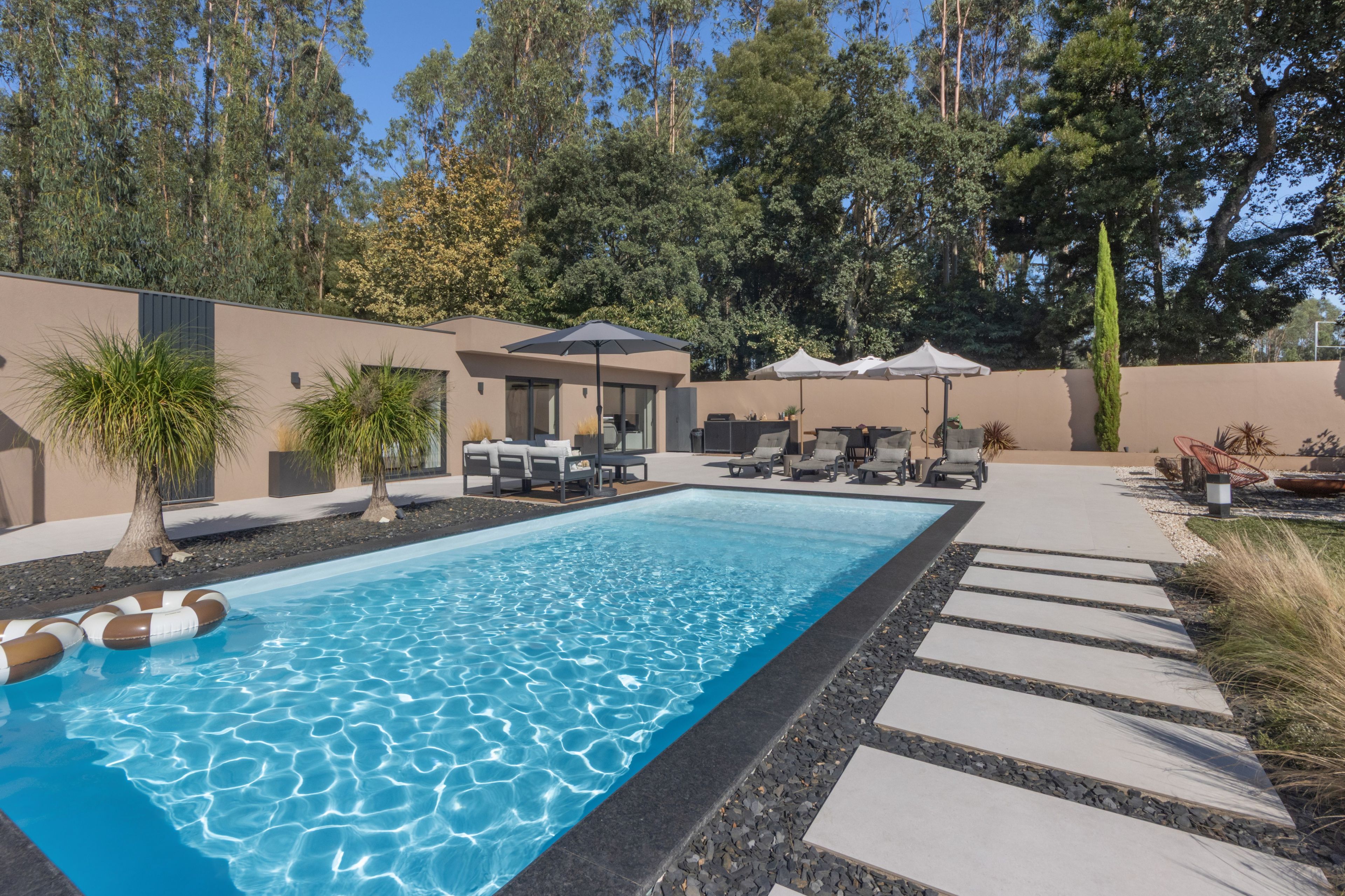 Ein Swimmingpool mit Liegestühlen und Sonnenschirmen, umgeben von einem angelegten Garten und einem modernen Gebäude.