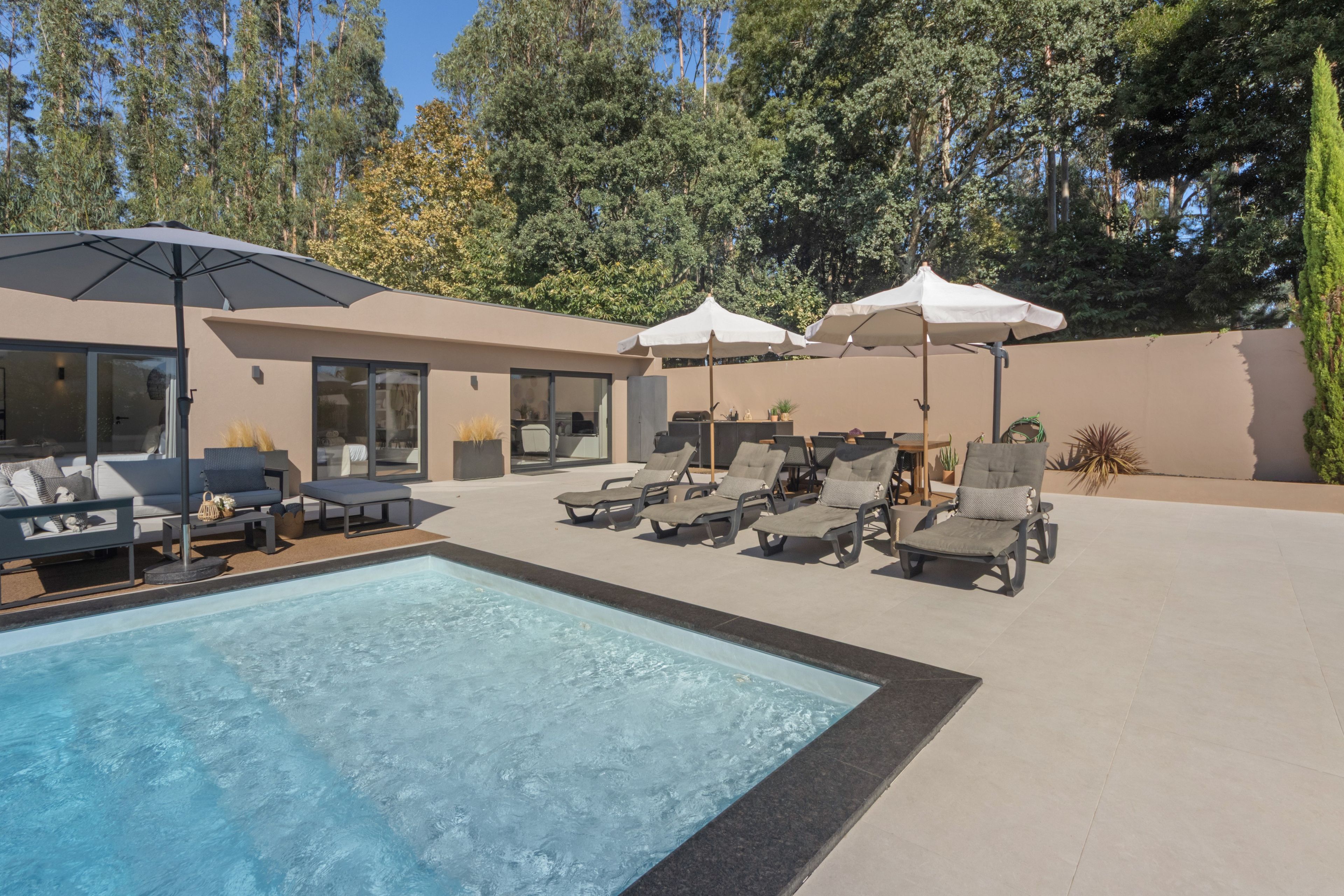Un espace piscine extérieur moderne, doté de chaises longues et de parasols, entouré d’un jardin aménagé et de grands arbres.