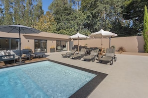 A modern outdoor pool area with lounge chairs and umbrellas, surrounded by a landscaped garden and tall trees.