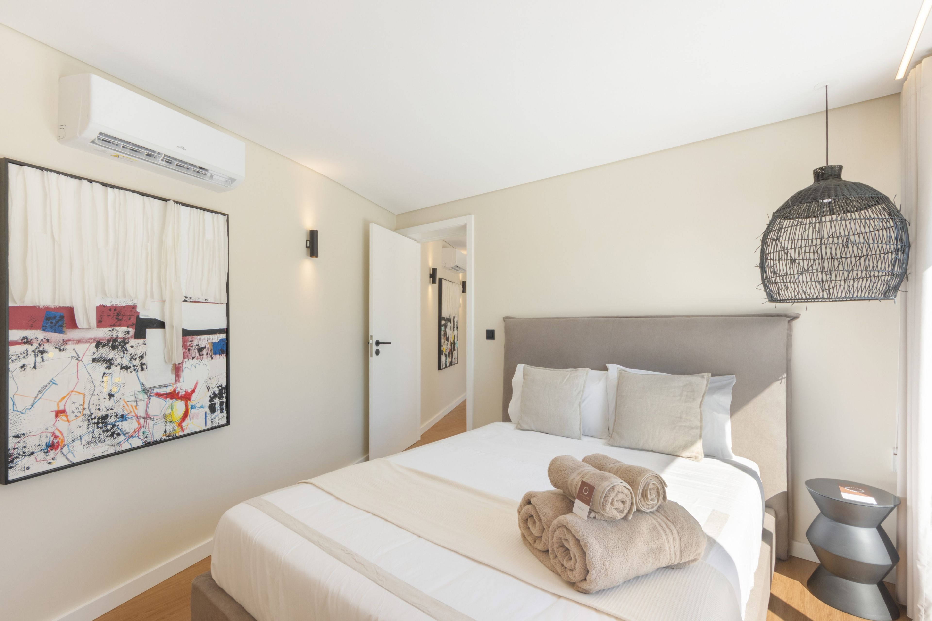 A hotel room with a bed, a wall-mounted air conditioner, a painting, and a hanging light fixture.