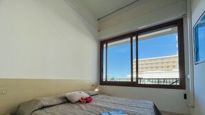 A bedroom with a large window, a bed with patterned bedding, a bedside table, and a nightstand. - Sea view apartment in central Bibione (Bibione)
