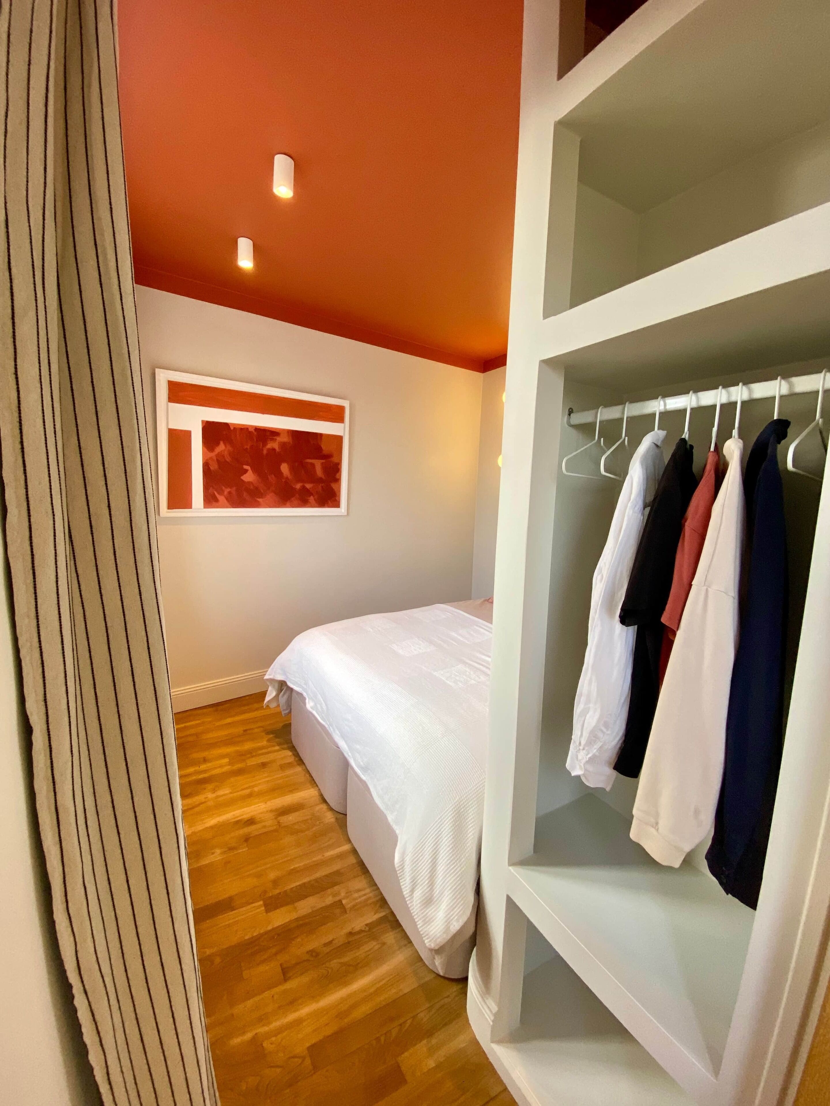 Ein Schlafzimmer mit einem Bett, einem Kleiderschrank mit Kleidung, einem Fenster mit Kunstwerk und einem Holzboden.