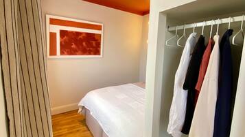 Ein Schlafzimmer mit einem Bett, einem Kleiderschrank mit Kleidung, einem Fenster mit Kunstwerk und einem Holzboden.