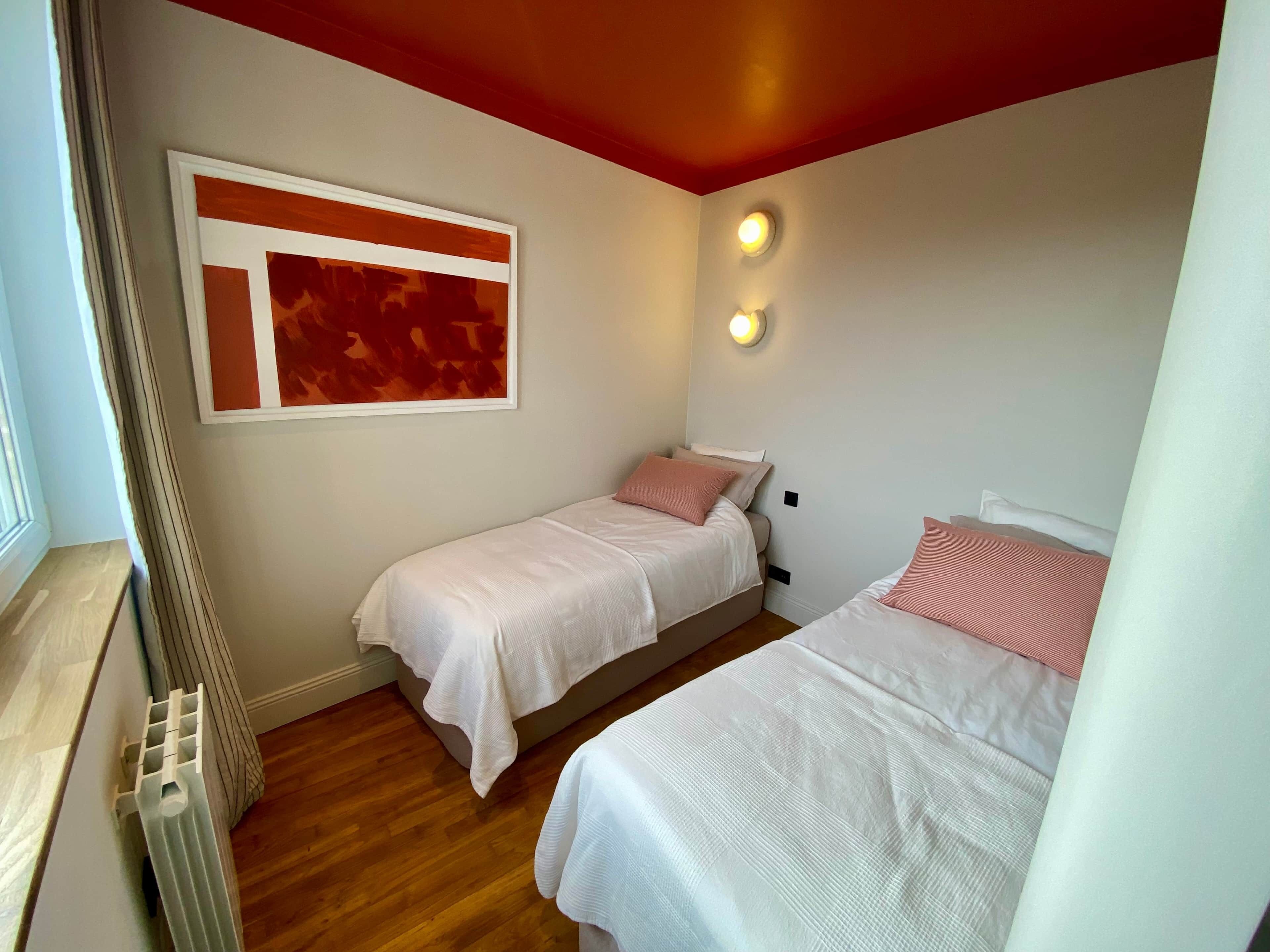 Une petite pièce bien éclairée, avec deux lits, un radiateur et un luminaire fixé au mur.