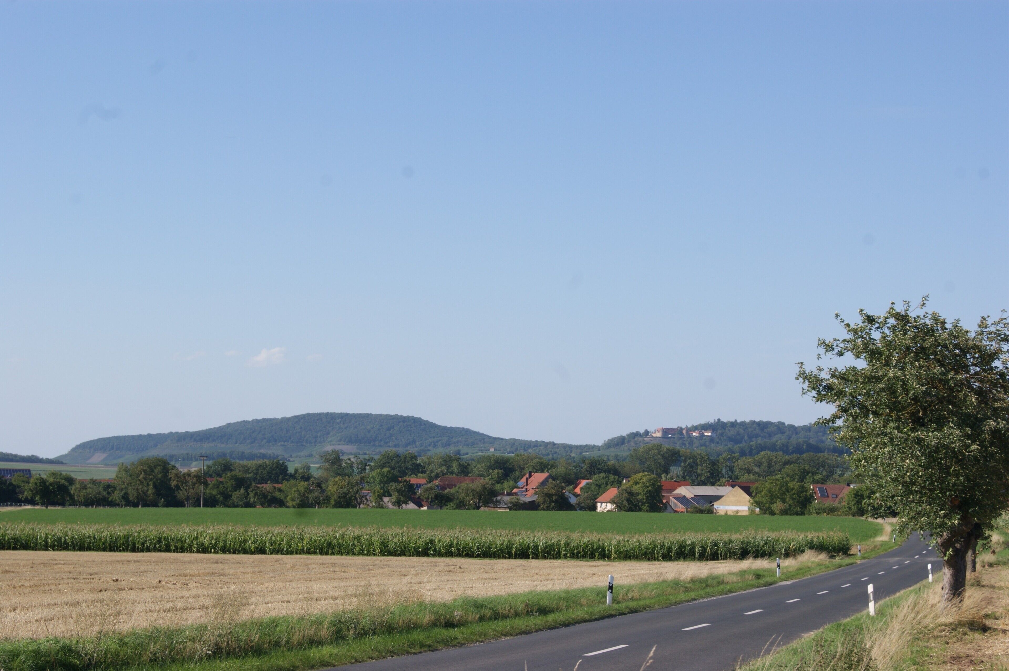 A rural road with houses and fields, a clear sky, and distant hills.