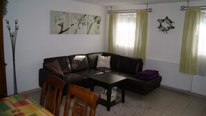 Un salon avec un canapé en cuir marron, une table à manger en bois et une table basse. Il y a deux fenêtres avec des rideaux, un radiateur et des œuvres d’art accrochées au mur.