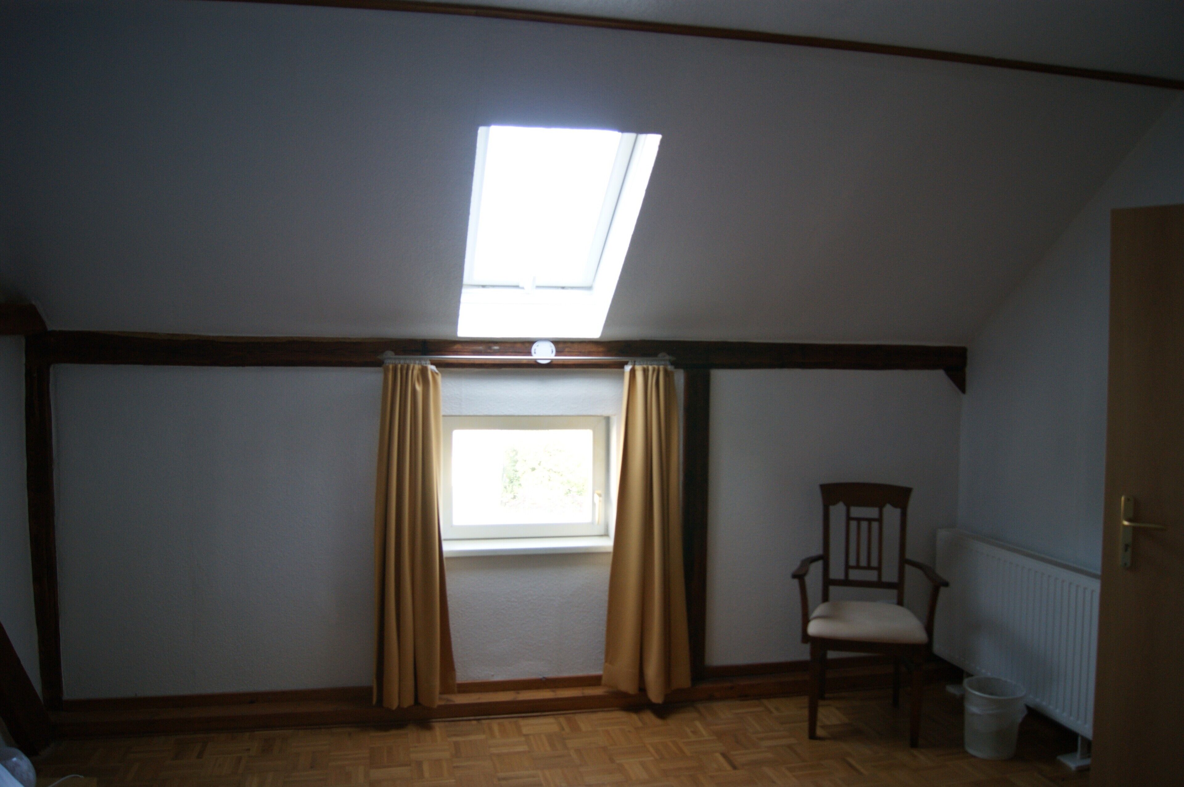 Una habitación con techo inclinado, un tragaluz, una ventana con cortinas, una silla de madera y un radiador.