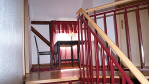 Un escalier avec une rampe en bois et des éléments décoratifs rouges, menant à une pièce où se trouvent une table et une chaise.