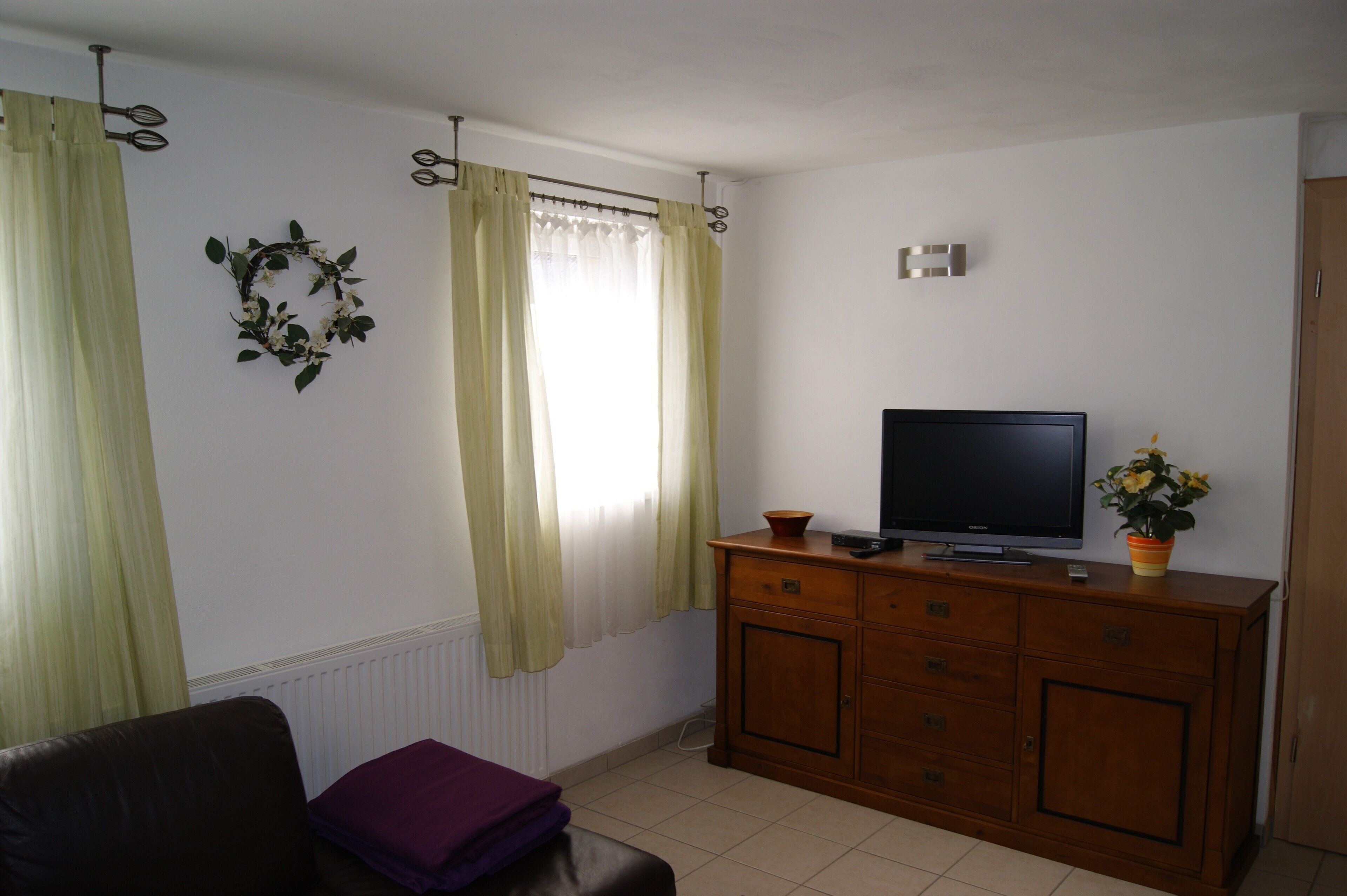 A living room with a television on a wooden cabinet, a window with curtains, a radiator, and a wall-mounted wreath.