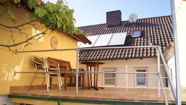 Un immeuble résidentiel avec un toit recouvert de tuiles, une antenne parabolique, une porte de garage verte et un petit balcon agrémenté d’une chaise et d’une table.