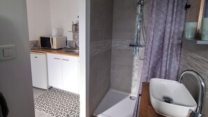 Un baño moderno con ducha, lavamanos y una pequeña zona de cocina al fondo.