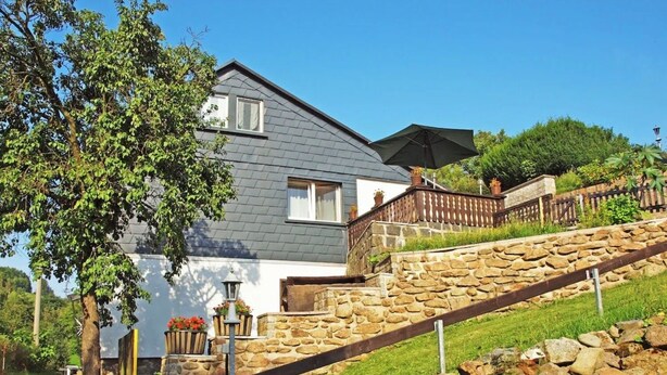 A two-story house with a stone staircase leading to a wooden deck, surrounded by greenery.