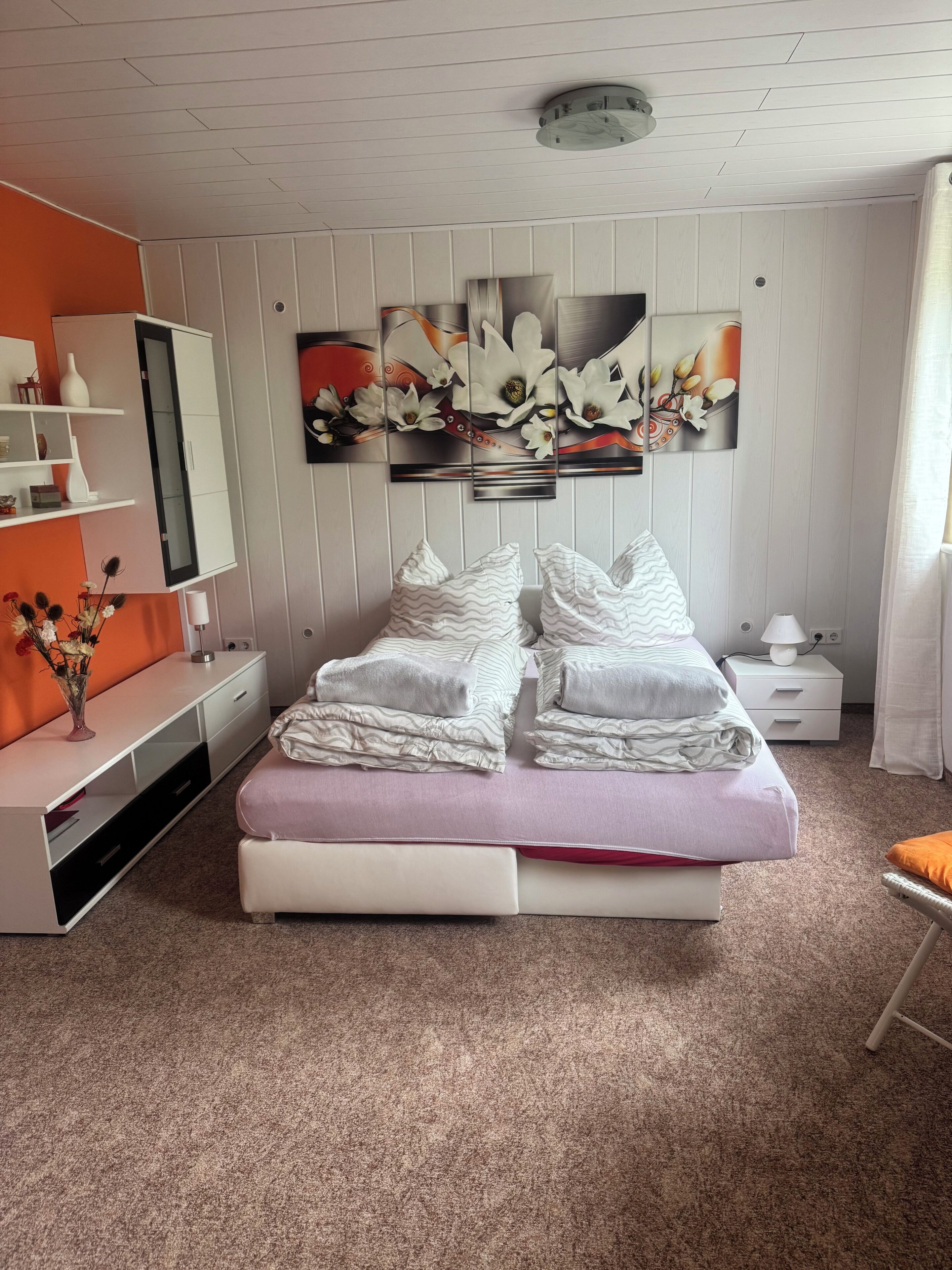Ein Schlafzimmer mit einem Bett, einem Nachttisch, einem Spiegel, einem Wandbild und einem Regal mit Dekoration.
