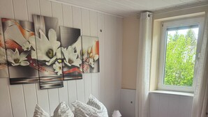 A neatly made bed with pillows and a blanket, a wall with floral artwork, a window with a view of greenery, and a radiator.
