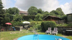 A backyard with a swimming pool, a wooden deck, and a fenced area with chairs and umbrellas.