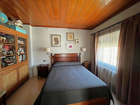 Un dormitorio con techo de madera, una cama con cubrecama gris, un aparador de madera, dos lámparas de noche y una ventana con cortinas.