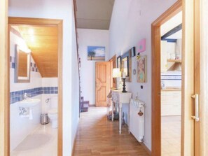 Un pasillo con un baño a la izquierda, un radiador y decoración en las paredes en el centro, y una zona de cocina a la derecha.