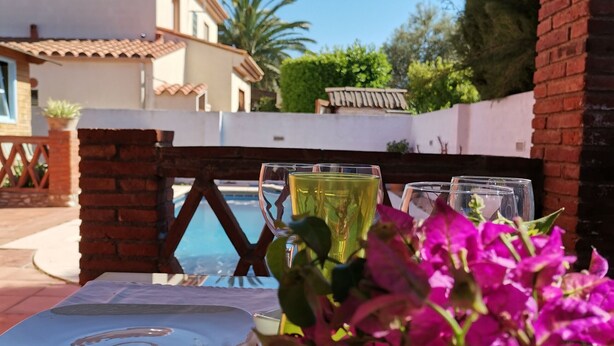 A table with a glass of yellow liquid, a clear glass, and a white plate, set against a backdrop of a swimming pool and a building with a tiled roof. - Holiday home with private pool, Sant Pere Pescador (Sant Pere Pescador)