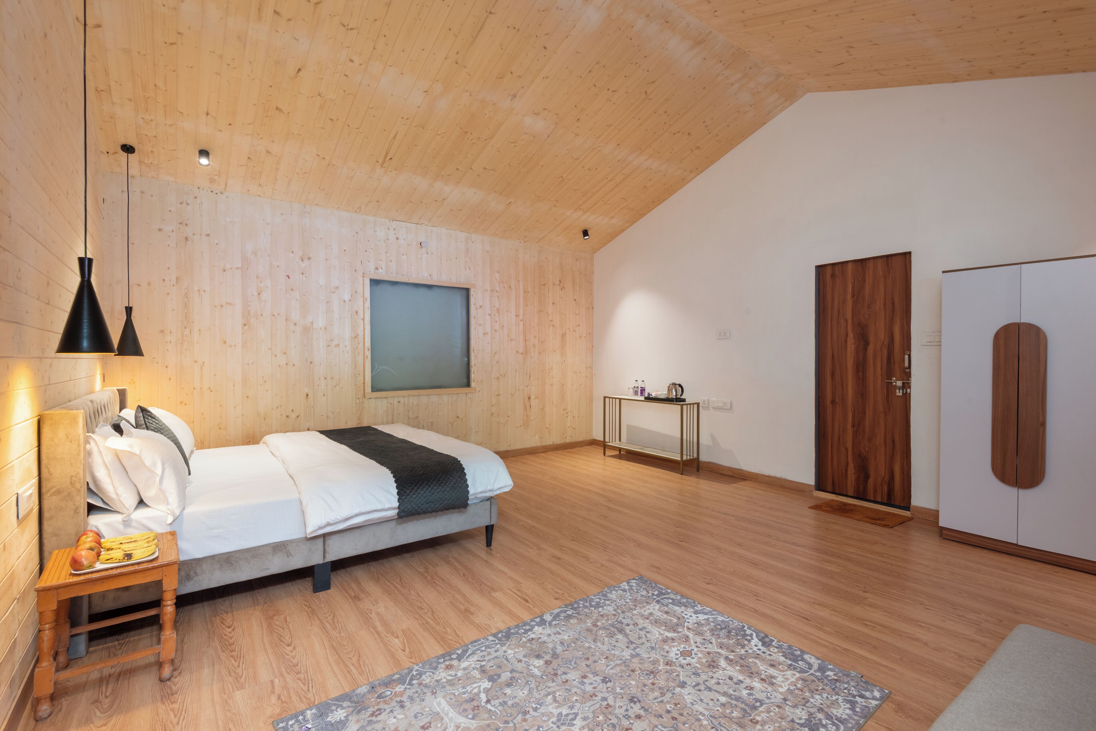 A bedroom with a wooden ceiling, a bed, a bedside table with a lamp, a small table with toiletries, a wardrobe, and a large window.