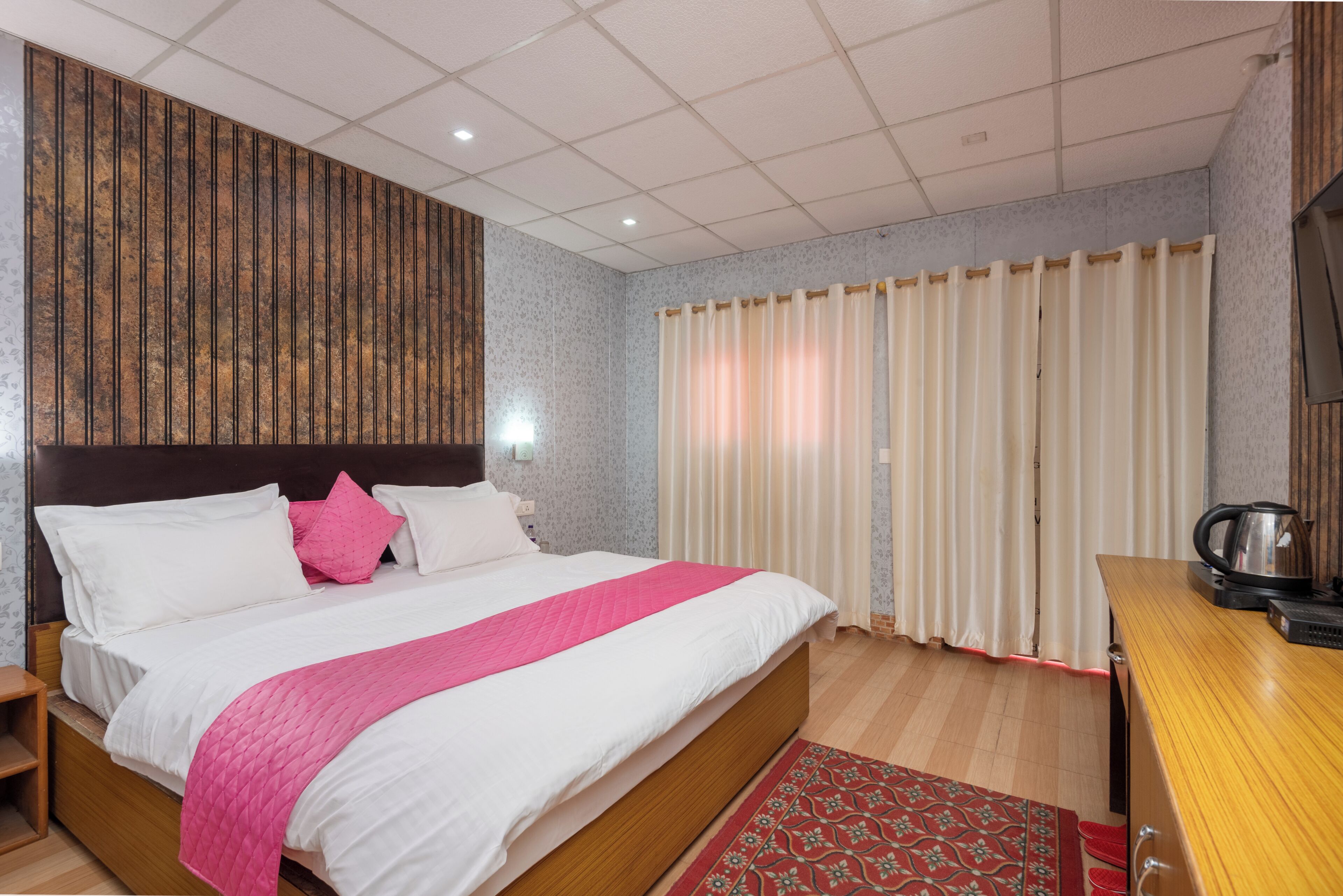 A hotel room with a bed, a wooden headboard, a nightstand, a desk, a kettle, and a window with curtains.
