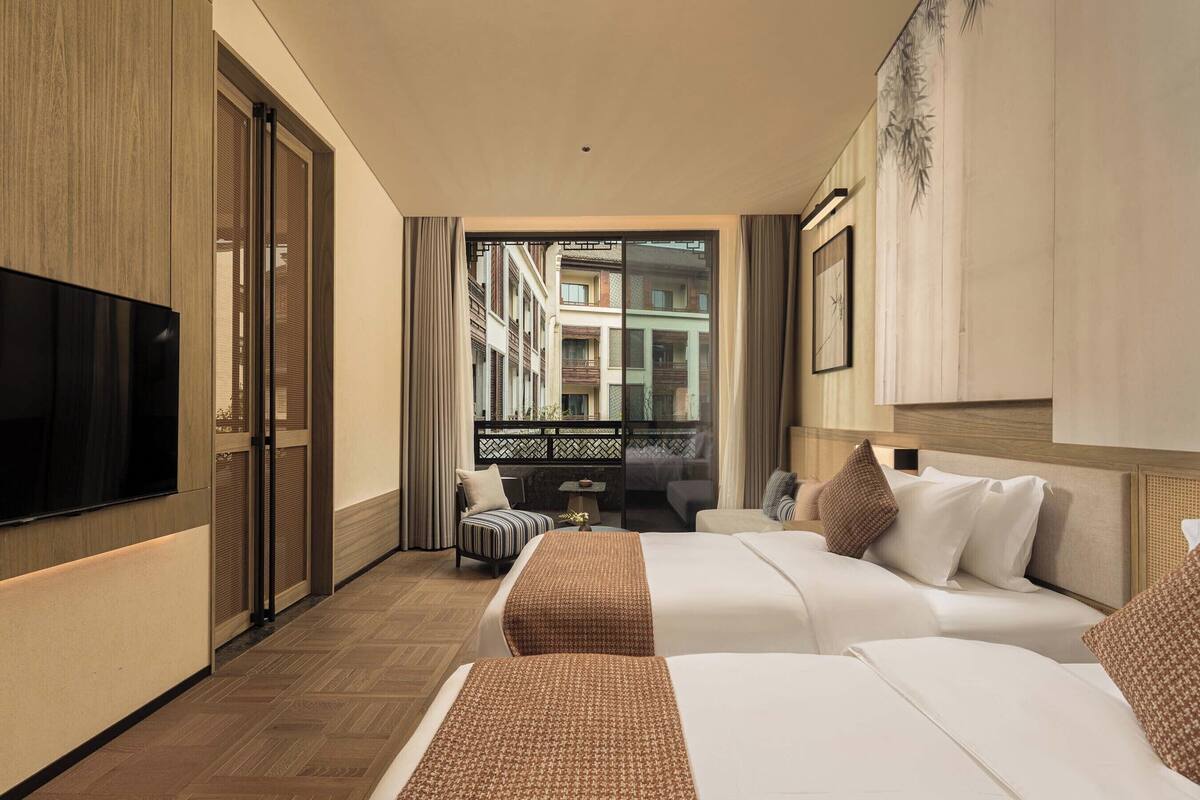 Ein Hotelzimmer mit zwei Betten, einem Fernseher, einem Balkon mit Blick auf Gebäude und einem Bild an der Wand.