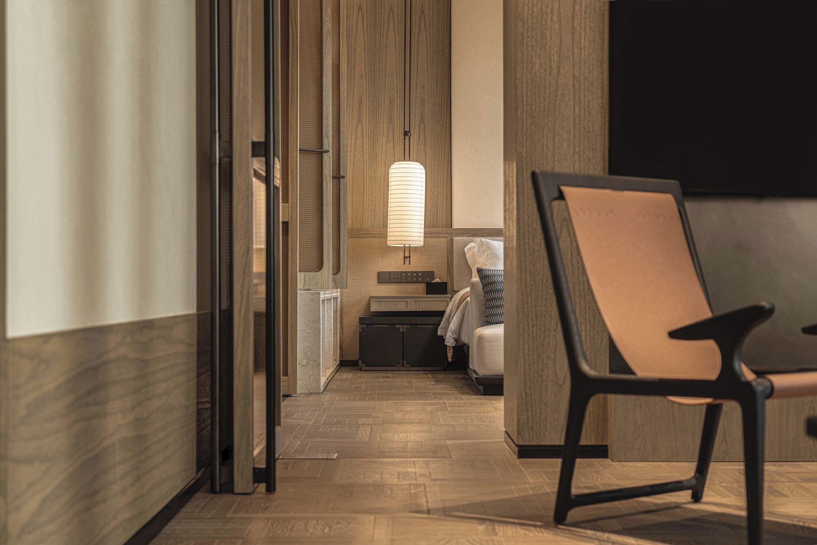 A hotel room with a wooden floor, a bed with a white headboard, a chair, a bedside table, and a wall-mounted TV.