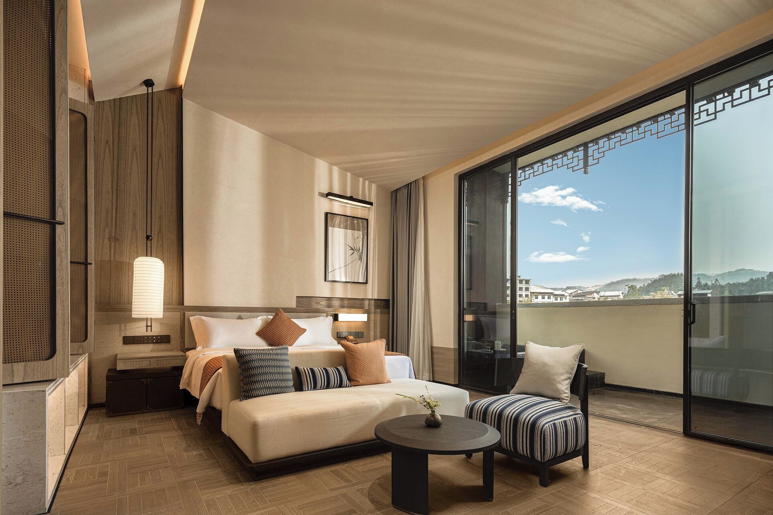 A modern hotel room with a large bed, a small sofa, a round coffee table, a nightstand, and a large window offering a city view.