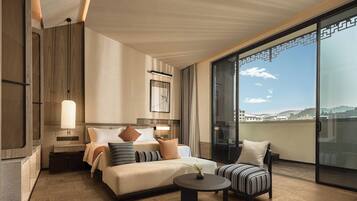 A modern hotel room with a large bed, a small sofa, a round coffee table, a nightstand, and a large window offering a city view.