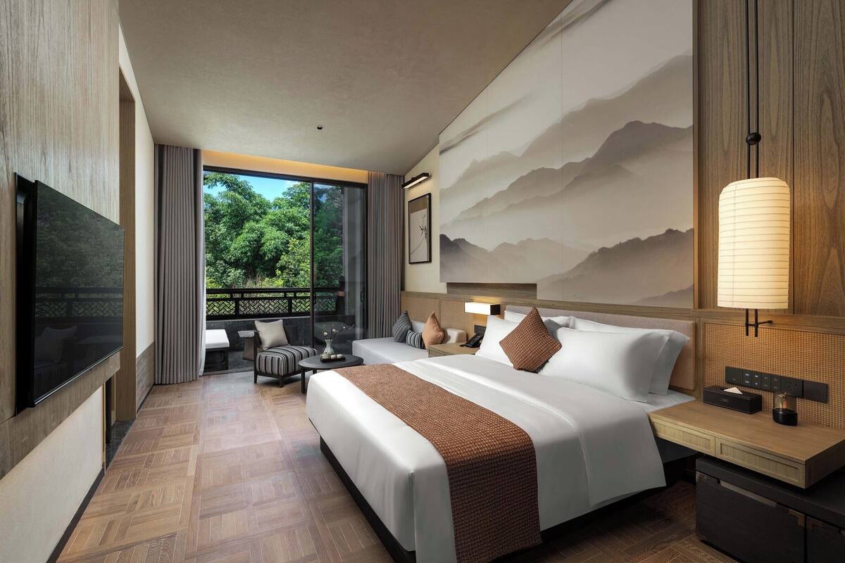 Ein Hotelzimmer mit einem großen Bett, einem Schreibtisch, einem Fernseher und einem Balkon mit Blick auf Bäume.