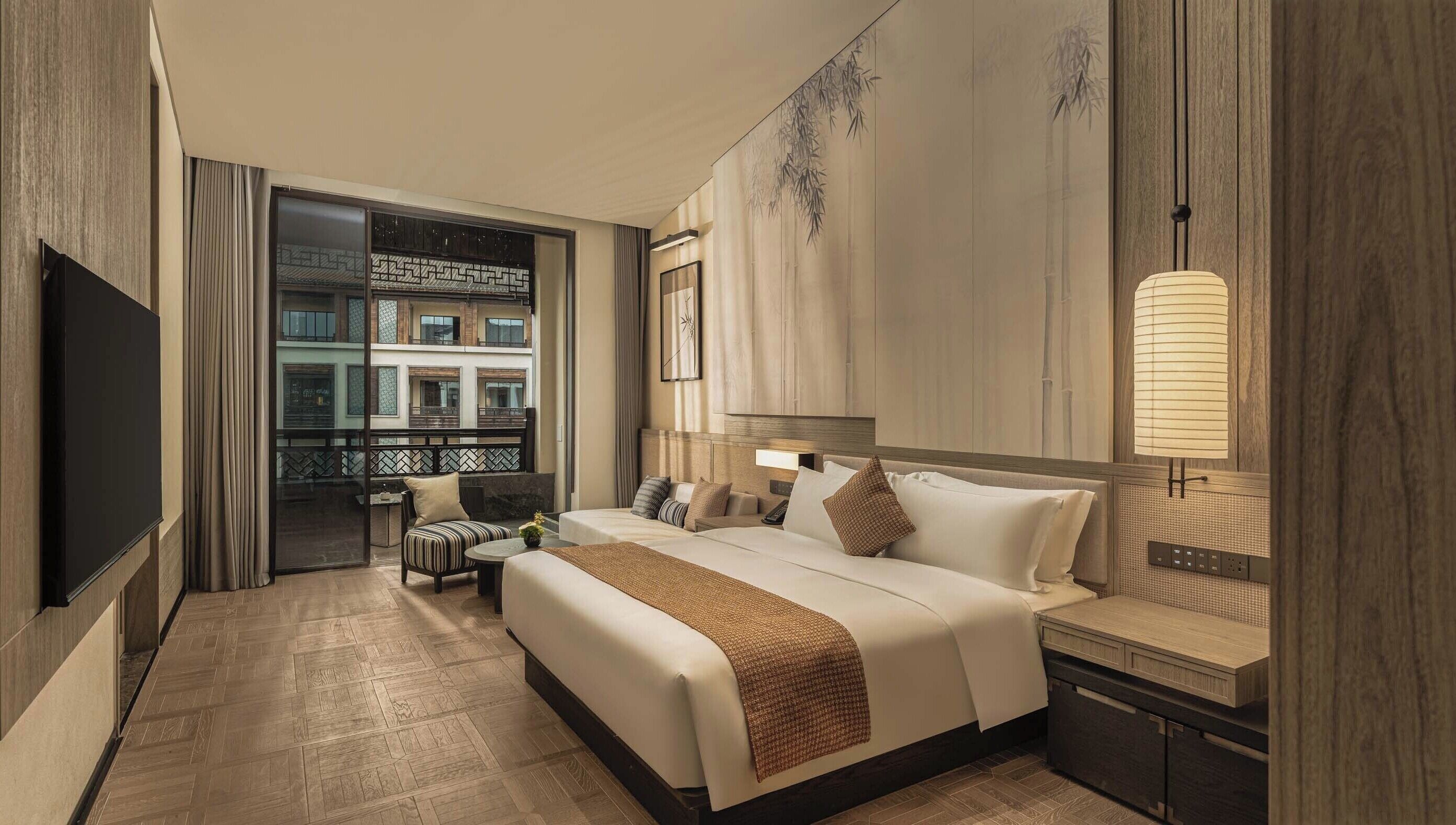 A hotel room with a bed, bedside table, lamp, and a view of a building through a glass door.