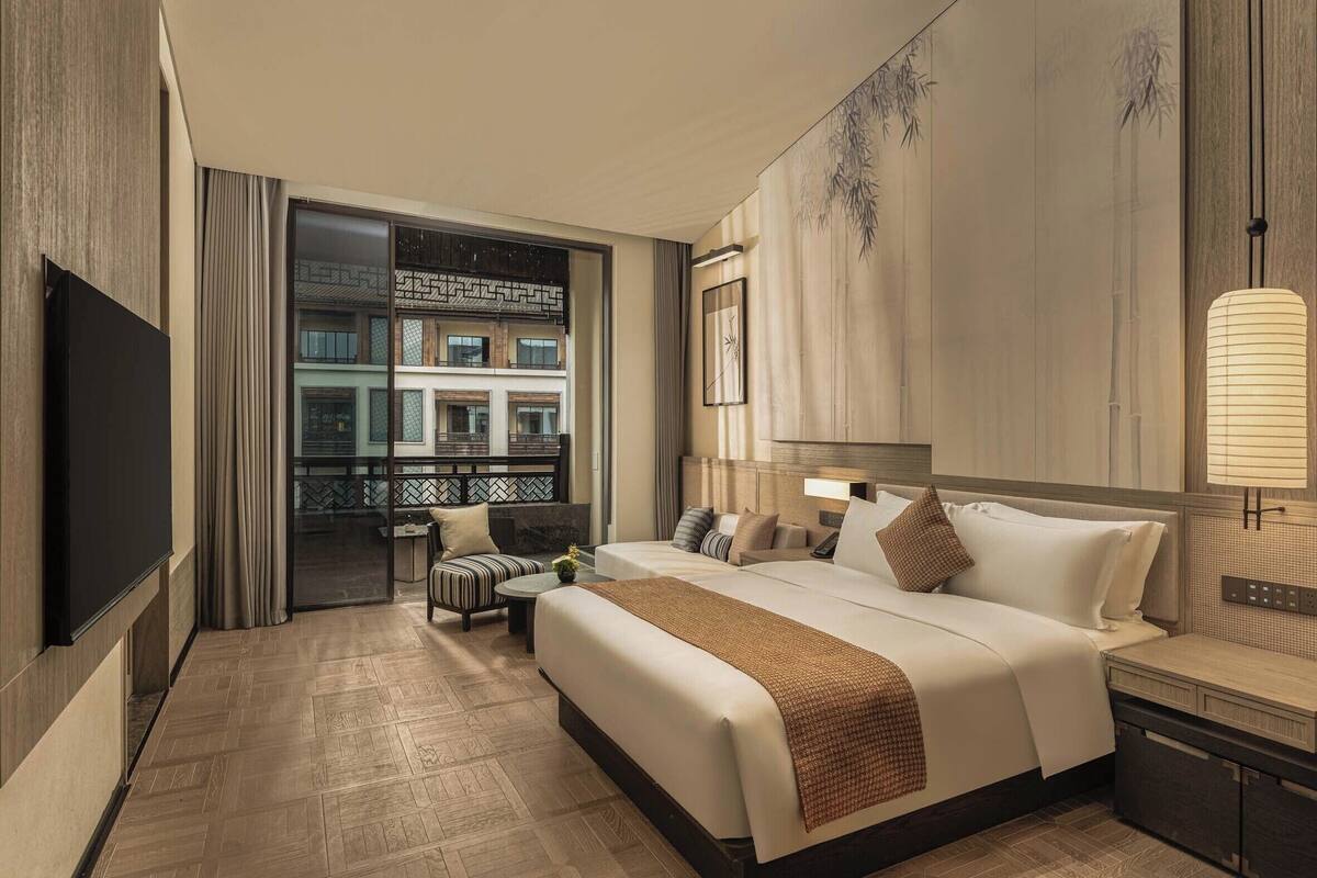 Ein Hotelzimmer mit Bett, Nachttisch, Lampe und Blick auf ein Gebäude durch eine Glastür.