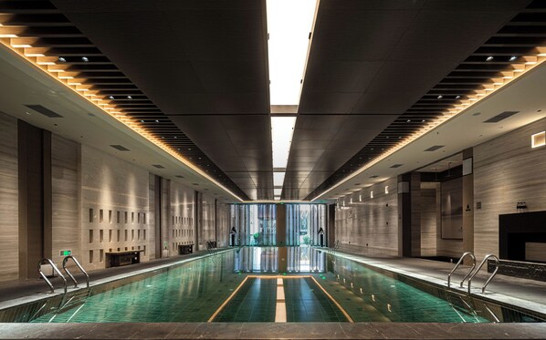 A modern indoor swimming pool with a central walkway, surrounded by walls with recessed lighting and a skylight.