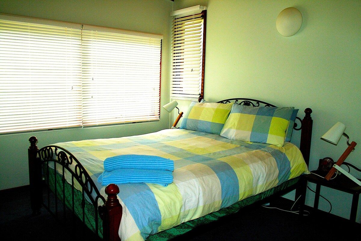 A bedroom with a bed, two windows, a bedside lamp, and a guitar on a nightstand.
