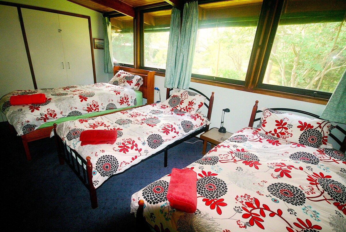 Ein Zimmer mit drei Einzelbetten, jeweils mit einem blumigen Überwurf und einem roten Kopfkissen. Es gibt einen Kleiderschrank und einen kleinen Nachttisch mit einer Lampe.