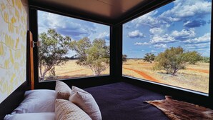 A room with a bed, pillows, a blanket, and a view of a dry landscape with trees and a dirt road.