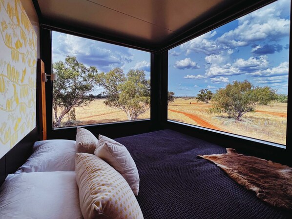A room with a bed, pillows, a blanket, and a view of a dry landscape with trees and a dirt road.