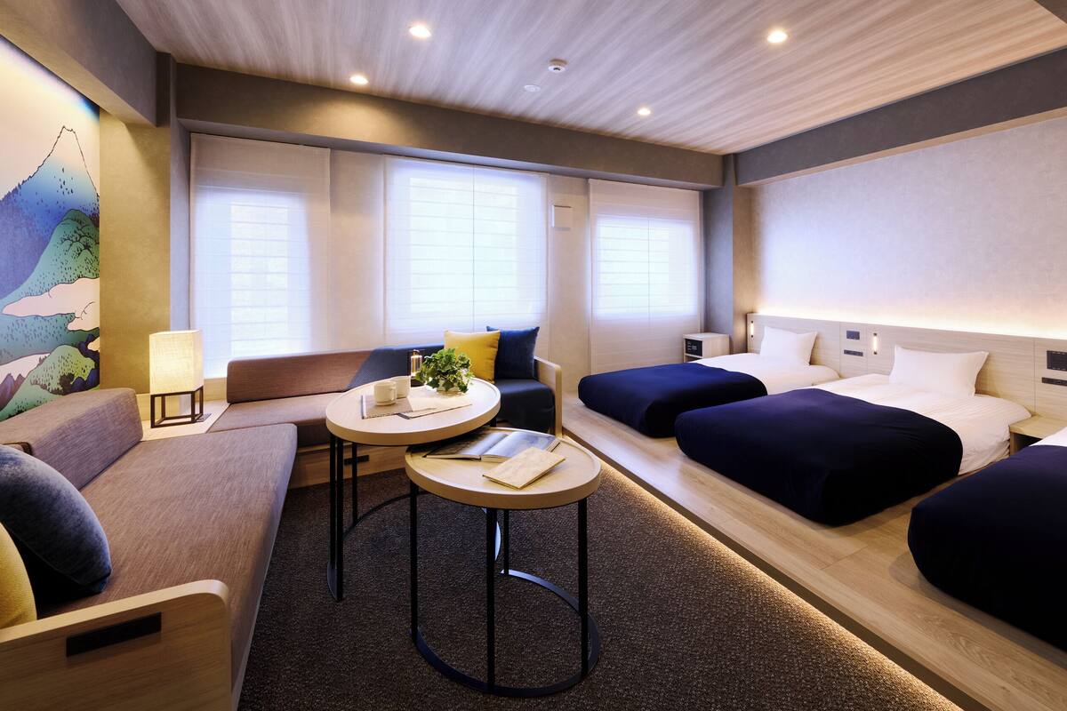 A hotel room with a sofa, two single beds, a small table with a lamp, and a painting on the wall.