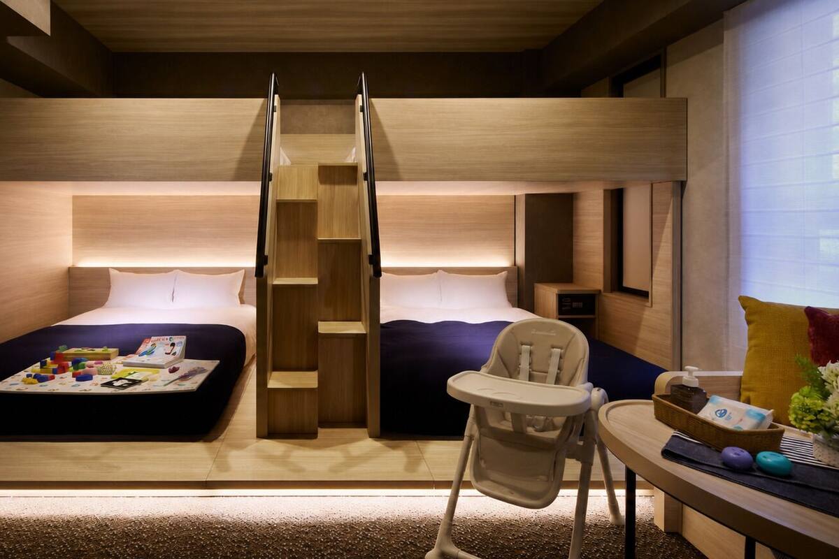 A compact hotel room with two beds, a small table, a chair, and a built-in ladder.