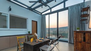 A modern room with a dining table, chairs, a wooden cabinet, and a large window with a view. - ELIVAAS White House 7BHK Villa  (Igatpuri)