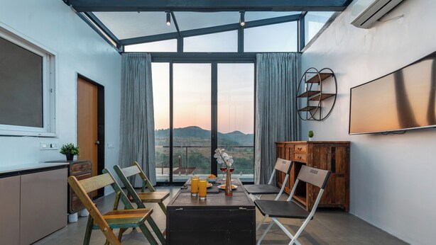 A modern room with a large window, a dining table with chairs, a TV, and a wooden cabinet. - ELIVAAS White House 7BHK Villa  (Igatpuri)