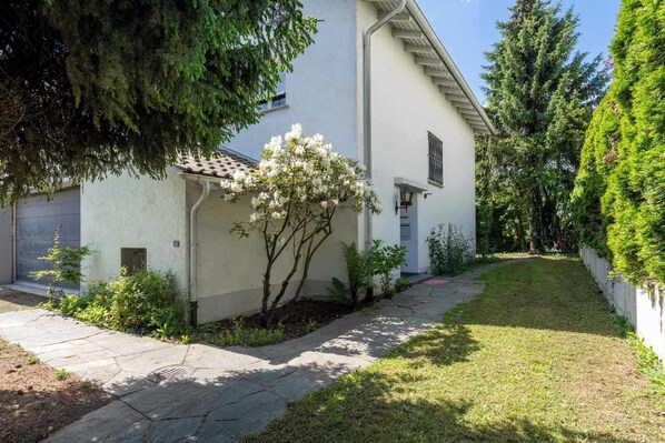 A white building with a tiled roof, a small garden with white flowering plants, and a paved walkway.
